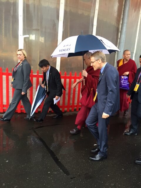 Dalai Lama walking under an umbrella with a group of people.