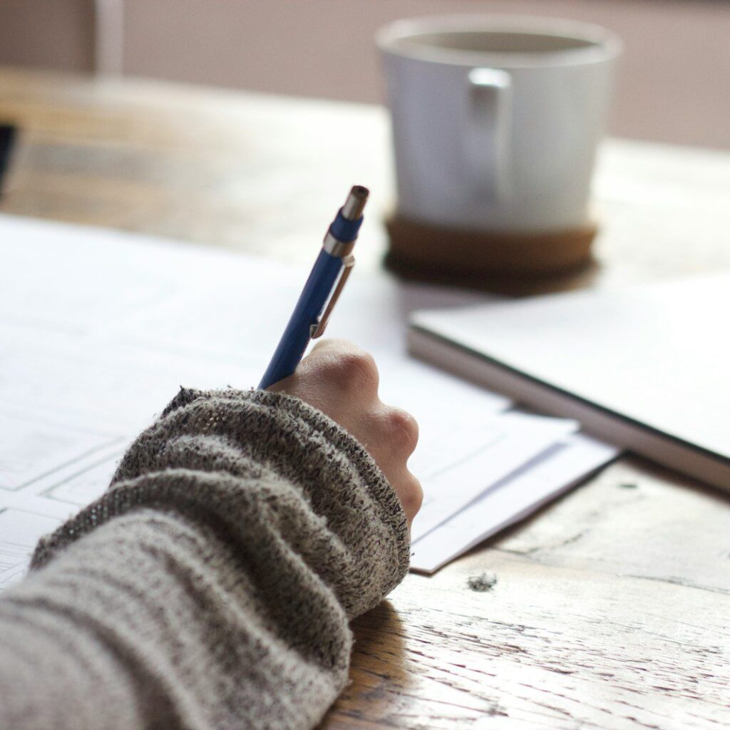 person writing on brown wooden table near white ceramic mug
