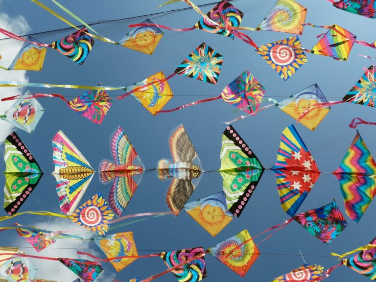 Festive, colorful kites shaped like diamonds and birds flying against a blue sky with a whisp of white cloud.