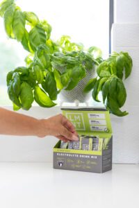 A hand is removing a packet of LMNT from a box on a white counter. A leafy potted plant sits on the windowsill above the counter.