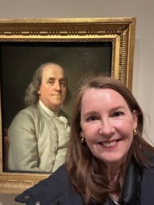A selfie of Gretchen smiling in front of a gold-framed portrait of Benjamin Franklin