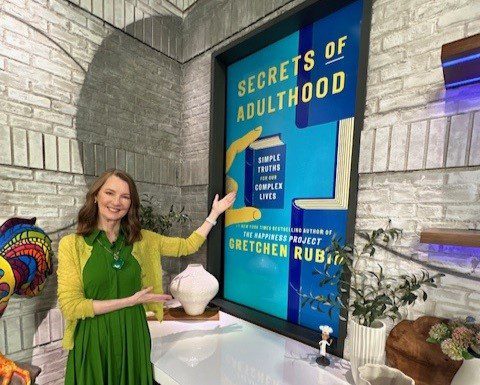 Gretchen poses next to a large poster showing the book cover of Secrets of Adulthood