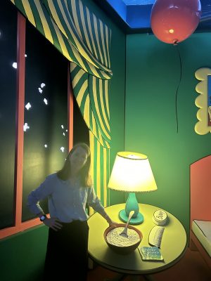 Gretchen in the great green room Gretchen smiling in a real-life version of a page from Goodnight Moon. Behind her is a window showing an illustrated night sky with a striped curtain falling across the corner. Next to her is a table holding an illuminated lamp and a bowl full of mush.